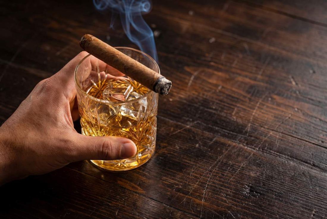 Hand holding a glass of whiskey with ice and a lit cigar on a dark wooden table
